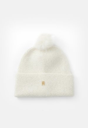 Bonnet tricoté blanc avec un motif diagonal texturé, comportant un pompon duveteux sur le dessus et un petit accent logo doré.