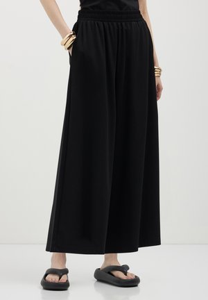 Pantaloni neri a gamba larga realizzati in tessuto morbido con vita elastica, dotati di tasche laterali e una finitura liscia.