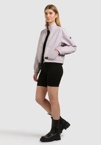 Jeune femme debout, portant une veste zippée gris clair, un short moulant noir et des bottines noires épaisses, sur un fond uni.