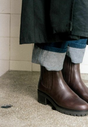 Bottines en cuir marron foncé avec des talons blocs, portées avec un jean bleu retroussé et un long manteau noir sur un sol texturé.