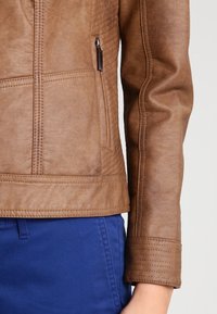 Bruine jas van nep leer met doorgestikte accenten, een ritszak en een gestructureerde pasvorm met een gladde textuur. Blauwe broek zichtbaar eronder.