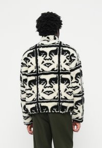 Fleecejakke i sort og hvid med et mønstret ansigt design. Tekstureret stof med en let oversized pasform og høj krave.