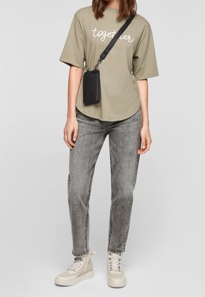 Persoon draagt een olijfgroen t-shirt met de tekst "together", grijs gewassen spijkerbroek, crèmekleurige hoge sneakers en een zwarte crossbody telefoontasje.