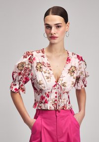 Blumenbluse mit Puffärmel, pinker Grundton mit roten und braunen Blumen, geraffter Kragen, gebundenes Vorderteil, kombiniert mit rosafarbene High-Waist-Hosen.