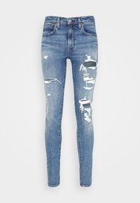 Jeans de mezclilla azul claro, desgastados y con detalles rasgados, diseño ajustado, herrajes dorados y estilo clásico de cinco bolsillos. La textura de la mezclilla es visible.