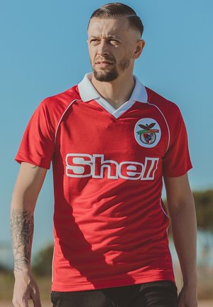 Rode voetbalshirt met witte kraag en het "Shell"-logo op de voorkant. Bevat een cirkelvormig embleem met een adelaar en tekst aan de zijkant.