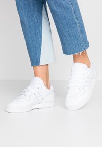 Sneakers bianchi in pelle, con dettagli perforati, tre righe laterali e una suola in gomma testurizzata. Indossati con pantaloni in denim a sigaretta.