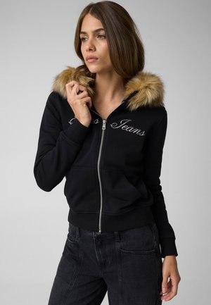 Ung kvinde iført en sort hættetrøje med lynlås og pelskant på hætten samt mørke jeans, kiggende til siden mod en ensfarvet baggrund.