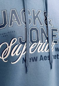 Μπλε φούτερ με κουκούλα από απαλό ύφασμα, διαθέτει μεγάλα λευκά και ναυτικά γράμματα και τελειώνει με πλεκτά κορδόνια.