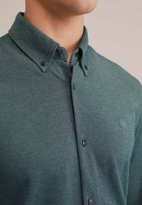 Temno zelena srajca s teksturirano tkanino, z razširjenim ovratnikom, črnimi gumbi in majhnim logotipom na prsih.