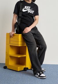 Svart Nike-t-shirt med vitt logo, grå jeans och svartvita sneakers. Sitter på en lysande gul förvaringsenhet med hjul.