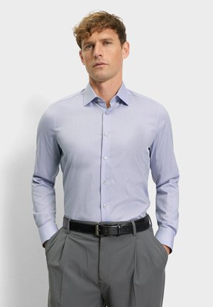 Mann trägt hellblaues Hemd, in graue Hose mit schwarzem Gürtel gesteckt, steht mit den Händen in den Taschen vor einfachem Hintergrund.