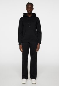 Svart hoodie med känguruficka, dragsko på huvan och ribbade ärmslut, kombinerad med matchande svarta sweatpants och vita sneakers.