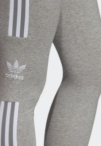 Leggings atletici grigi realizzati in tessuto morbido, con tre strisce verticali bianche e un logo ricamato vicino alla coscia.