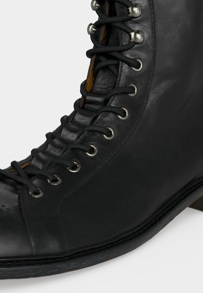 Μαύρο δερμάτινο μποτάκι με κορδόνια, μεταλλικά ρούχοι και ενισχυμένη μυτερή μύτη, εμφανίζεται σε κοντινή πλαϊνή λήψη σε απλό φόντο.