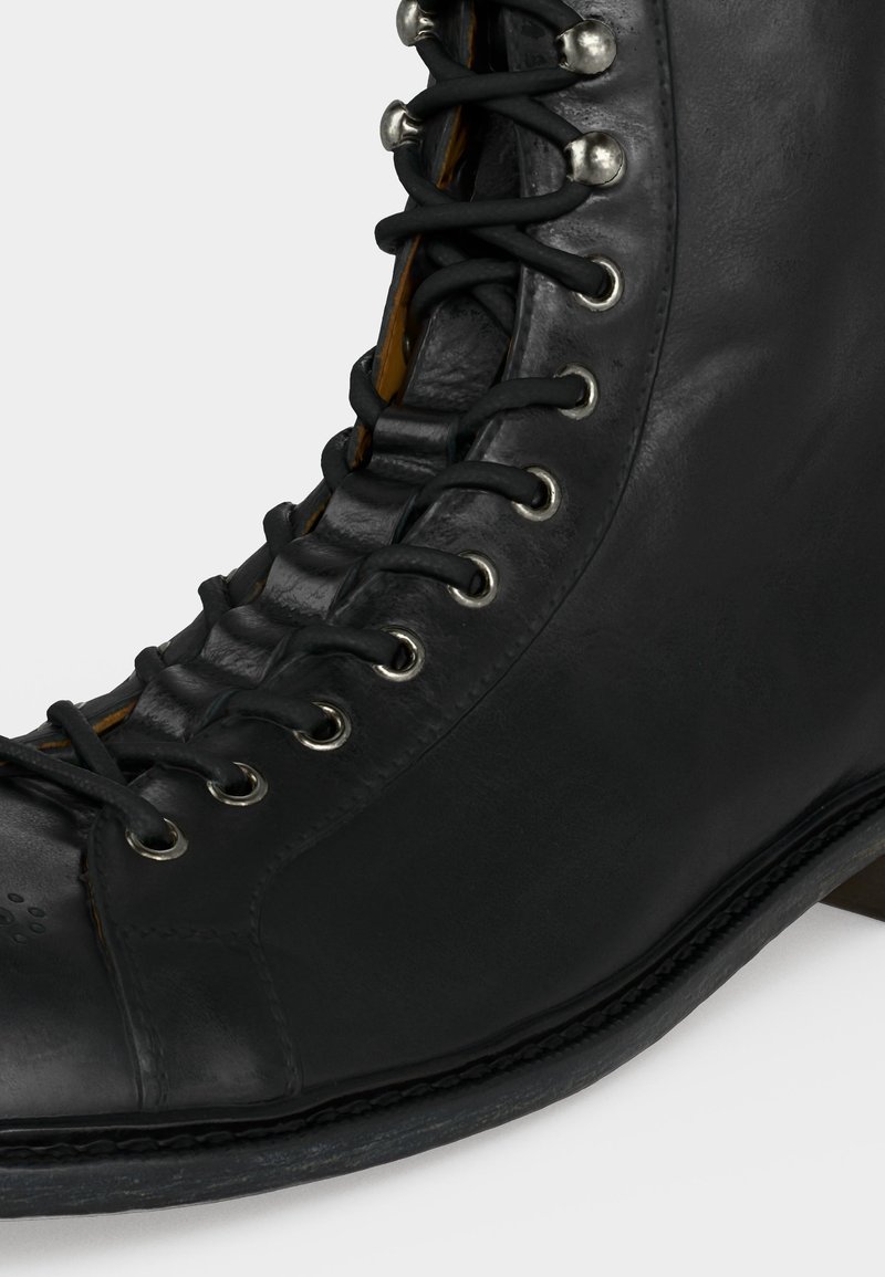 Schwarzer Leder-Schnürstiefel mit Metallösen und verstärkter Zehe, in Nahaufnahme seitlich vor einfachem Hintergrund gezeigt.