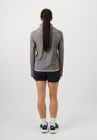 Šedý športový top s dlhým rukávom a kapucňou, ktorý má kontrastné švy. Skombinovaný s čiernymi šortkami a ponožkami; celkový dizajn je minimalistický.