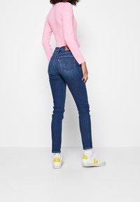 Vaqueros ajustados de denim azul oscuro, con bolsillos traseros visibles y un parche de cuero de la marca. Combinados con una camiseta entallada de color rosa claro y zapatillas blancas con detalles amarillos.