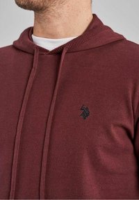 Sweat à capuche bordeaux en tissu doux avec une texture tricotée, doté d'un logo noir brodé sur le côté gauche de la poitrine et de cordons de serrage sur la capuche.