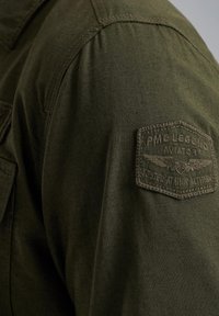Camisa de algodón verde oliva con un parche en el hombro izquierdo con texto en relieve y diseño de alas, tela texturizada y corte entallado.