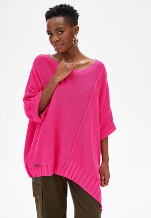 Kobieta w jaskrawo różowym, oversize, dzianym swetrze z asymetrycznym dołem i podwiniętymi rękawami, w połączeniu z oliwkowymi spodniami typu cargo i kolczykami kołami.