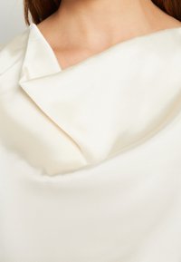 Kremowa satynowa bluzka z dekoltem w drapowanym, złożonym fasonie, gładką teksturą i subtelnym połyskiem. Brak widocznych elementów metalowych lub wzorów.