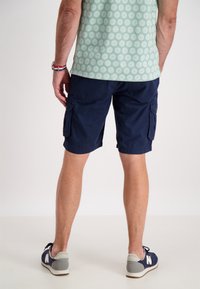 Marina cargo shorts med två sidfickor, tillverkade av ett lätt material. Bärs med en mönstrad grön skjorta och marinvita skor.