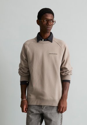Homme portant un sweat-shirt beige par-dessus une chemise noire à col, jean gris et lunettes, debout sur un fond gris clair.