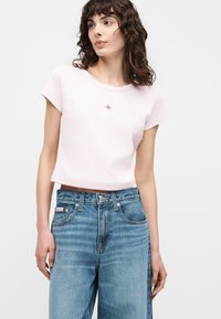 Γυναίκα που φοράει ροζ ανοιχτό cropped μπλουζάκι με μικρό λογότυπο και ψηλόμεσο μπλε τζιν, στέκεται μπροστά σε απλό φόντο.