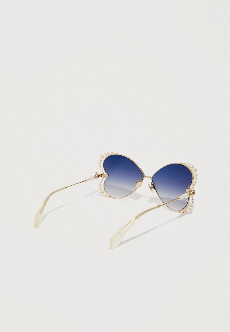 Lunettes en forme de cœur avec des verres dégradés bleus, monture dorée et accents en perles le long des bords. Branches transparentes et courbées.