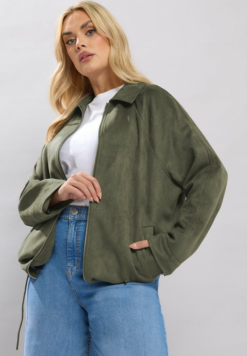 Donna bionda con giacca verde oliva non chiusa sopra una maglietta bianca e jeans blu, posa con una mano in tasca su sfondo grigio.