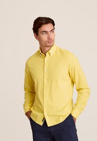 Geel overhemd met knoopjes, gemaakt van lichtgewicht stof, met een kraag en subtiele logo-details, gedragen met donkere broek tegen een neutrale achtergrond.