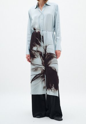 Vrouw die een lange lichtblauwe overhemdjurk draagt met zwarte abstracte bloemenprint, gestrikt in de taille, over wijde zwarte broek en schoenen.