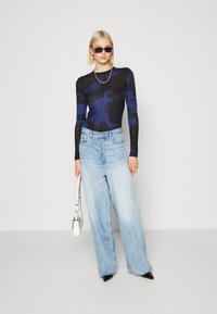 Maglietta a maniche lunghe con un motivo astratto blu scuro e nero, abbinata a jeans a gamba larga di un blu chiaro. Gli accessori includono piccoli occhiali da sole neri e una borsa bianca.