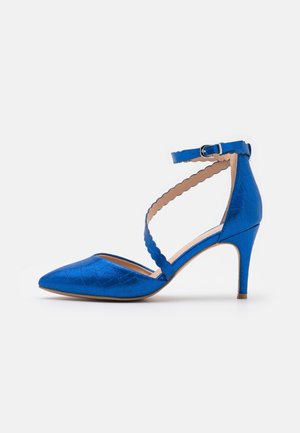 Chaussure à talons hauts bleu, texturée, avec un bout pointu, une bride de cheville et des détails en forme de vagues. Elle possède un talon fin de style stiletto.