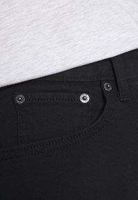 Svarta denimjeans med en framficka, metallnitar och en sl ät yta, med en grå tygöverdrag delvis synlig ovanför.