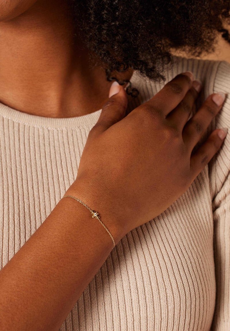 Gouden kettingarmband met een klein bloemontwerp, gedragen om een pols tegen een licht geribbeld topje. De armband heeft delicate schakels en een subtiele glans.
