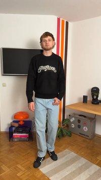 Juodas megztinis su "PEGADOR" tekstu, šviesiai mėlynos džinsai ir juodi sportbačiai. Stovi ant dryžuoto kilimo prieš spalvingą sieną.