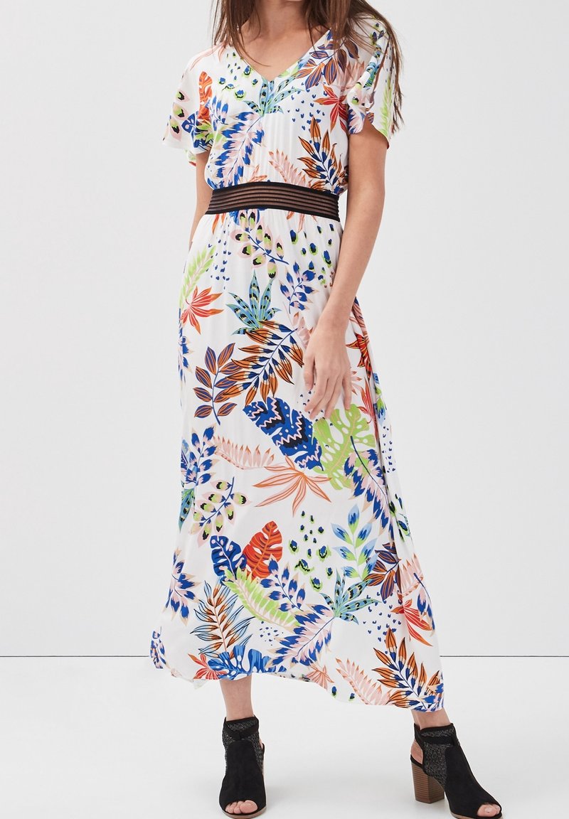 Femme portant une longue robe blanche à imprimé feuilles tropicales multicolores, ceinturée à la taille d'une ceinture noire rayée, associée à des talons blocs noirs ouverts à l'avant.