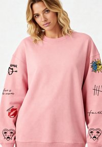 Roza majica s kapuljačom s naglašenim ramenima, rebrastim manžetama i rubom. Ukrašena raznim grafičkim motivima i tekstom duž rukava. Ležeran, širok kroj.