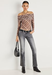 Γυναίκα που φοράει καφέ καρό τοπ έξωμο, ξεθωριασμένο γκρι τζιν, μαύρη ζώνη, μαύρα μυτερά μποτάκια, ασημένιο κολιέ, κρατώντας μαύρη τσάντα χειρός.