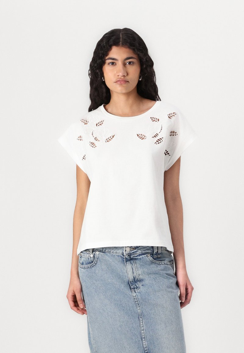 T-shirt de algodão branco com mangas curtas, apresentando padrões recortados de flores e folhas na frente. Combinado com uma saia de ganga azul.