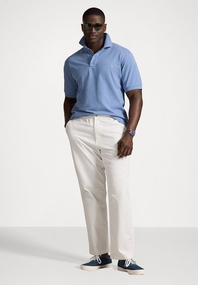 Camisa polo azul clara com colarinho, calças bege e sapatilhas azuis. Modelo a usar óculos de sol; relógio visível com mostrador azul.