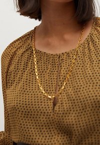 Brązowa bluzka w czarne kropki, z okrągłym dekoltem i małym otworem w kształcie klucza z przodu. Widoczny złoty naszyjnik.