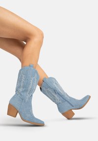 Niebieskie dżinsowe kowbojki z zakrzywionym noskiem, ozdobnym białym przeszyciem i piętką z klejonego drewna. Zamek błyskawiczny z tyłu.