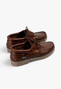 Zapatos náuticos de cuero marrón con punta redondeada, cordones de cuero y suela de goma texturizada. Presentan costuras blancas y un logotipo en el lateral.