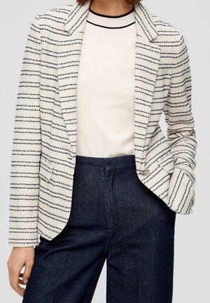 Žena oblečená v bílém saku s tenkými černými vodorovnými pruhy, bílém topu s černým lemováním u límce a tmavě modrých kalhotách.