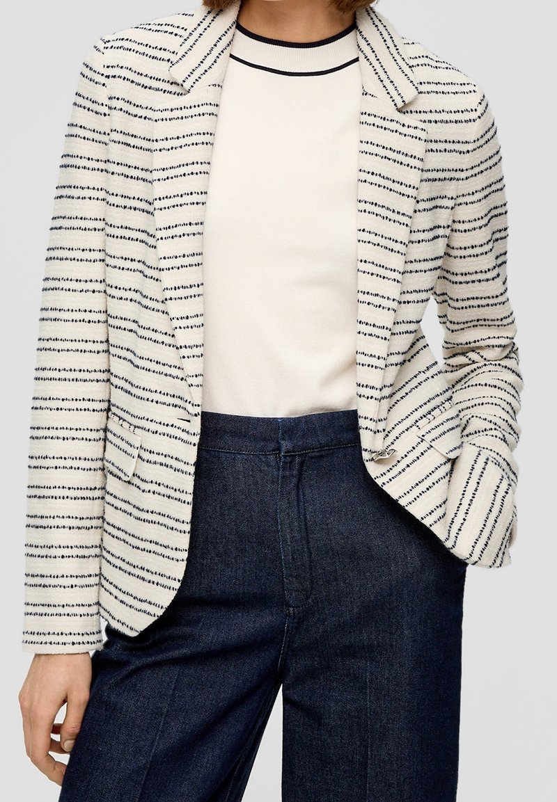 Kobieta ubrana w biały żakiet z cienkimi czarnymi poziomymi paskami, biały top z czarną lamówką przy kołnierzu oraz ciemnoniebieskie spodnie.