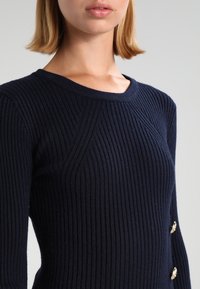 Marineblauer Rippstrickpullover mit Rundhalsausschnitt, strukturiertem Muster und goldfarbenen Knopfakzenten an der Seite.