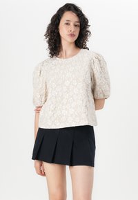 Kortermet, kremfarget bluse med blomsterdesign og puffede ermer, sammen med et svart plisserte skjørt. Stoffet har en teksturert overflate.